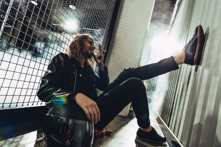 young man in leather jacket holding cigarette and helmet while sitting near gridの写真素材