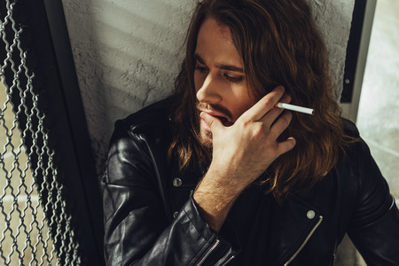 bearded long haired man in leather jacket smoking cigarette and looking awayの写真素材