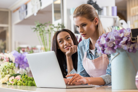 blonde florist in apron using laptop while pensive african american colleague looking away behindの写真素材