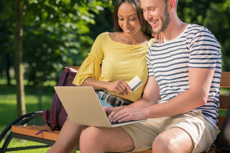 multiethnic couple shopping online with laptop and credit card while sitting on bench in parkの写真素材