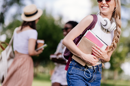 smiling blonde student with headphones holding books while friends talking behind in parkの写真素材