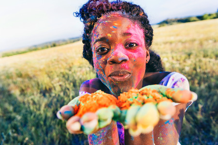 young african american woman blowing colorful powder from palms and looking at camera at holi festivalの写真素材