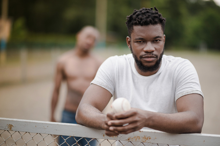 handsome african american man with baseball ball leaning on fence on courtの写真素材