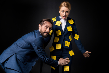 excited bearded businessman touching frustrated businesswoman with sticky notes on clothes looking at cameraの写真素材