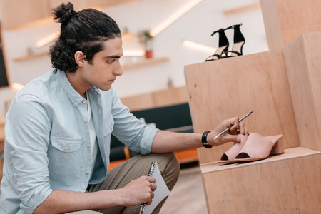 young man holding notebook with pencil while looking at shoes and working in boutiqueの写真素材
