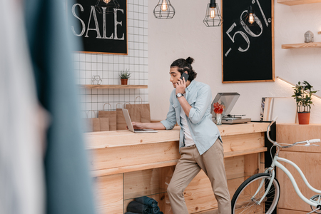 young boutique owner talking on smartphone and using laptop indoorsの写真素材