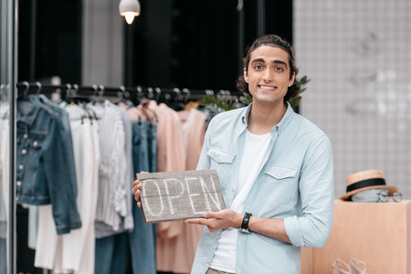 young shop owner holding open sign and smiling at cameraの写真素材
