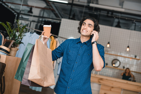 young man holding shopping bags and paper cup while talking on smartphone in boutiqueの写真素材