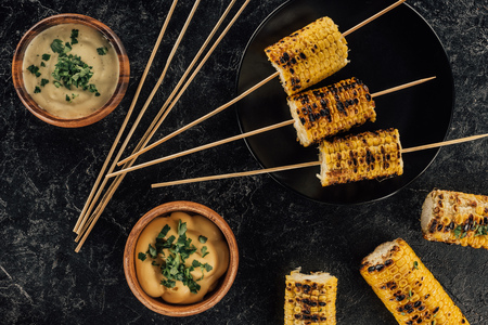 top view of grilled corn pierced with sticks and bowls of sauceの写真素材