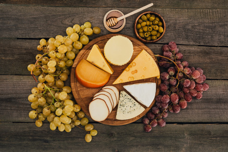 flat lay of assortment of cheese types on cutting board, grapes, honey and olives on wooden surfaceの写真素材