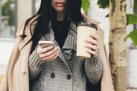 cropped view of woman in coat holding smartphone and coffee to goの写真素材
