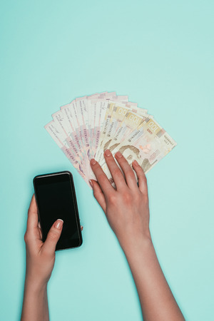 cropped shot of woman with cash and smartphone isolated on turquoise
