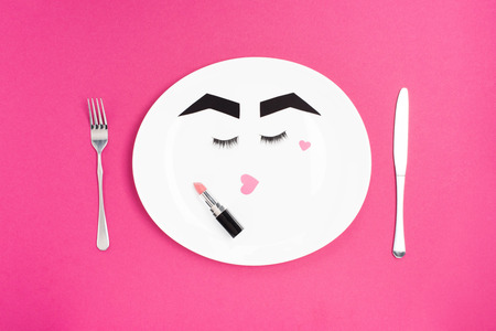 paper face with makeup on plate with fork, knife and lipstick isolated on pink, beauty conceptの写真素材