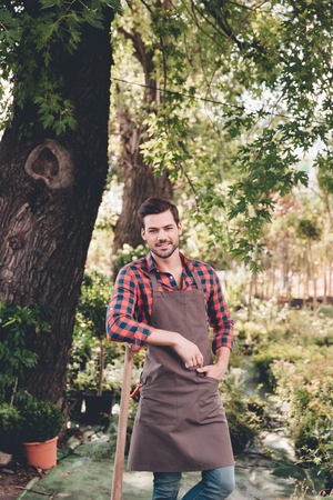 smiling gardener with hand in pocket looking at camera while standing in gardenの写真素材