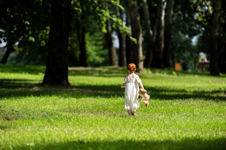 back view of cute little girl in white dress holding teddy bear and walking on green grass at parkの写真素材