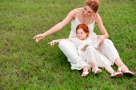 beautiful red haired mother and daughter reading book and pointing away with fingers on grass