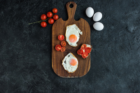 top view of arrangement of fried eggs and cherry tomatoes on wooden cutting board on dark surfaceの写真素材