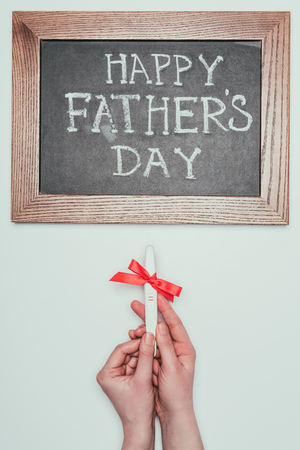 cropped shot of woman holding pregnancy test with ribbon and happy fathers day lettering on blackboardの写真素材