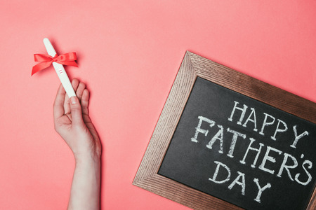 cropped shot of woman holding pregnancy test with ribbon and happy fathers day lettering on...