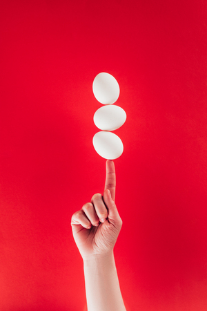 partial view of woman holding levitating raw chicken eggs on finger isolated on redの写真素材