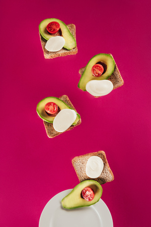 close up view of toasts, avocado pieces, mozzarella cheese and cherry tomatoes falling on plate isolated on pinkの写真素材