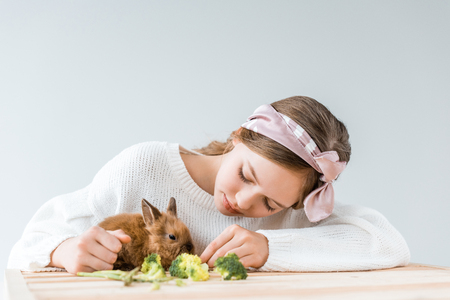 girl feeding cute furry rabbit with broccoli at wooden tableの写真素材