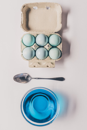top view of glass with blue paint, spoon and chicken eggs in trayの写真素材