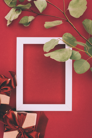 top view of empty white frame, gift boxes and twig with green leaves on redの写真素材