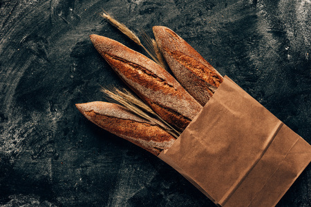 top view of arranged french baguettes in paper bag and wheat on dark tabletop with flour