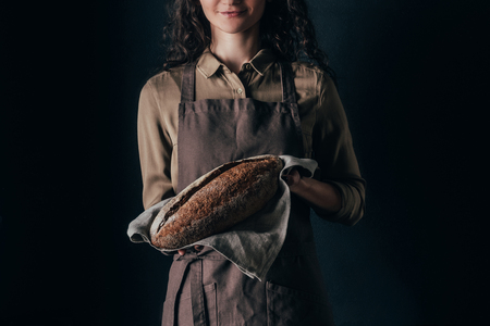 partial view of woman in apron holding french baguette in hands isolated on blackの写真素材