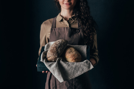 cropped shot of woman holding wooden box with loafs of bread in hands isolated on blackの写真素材
