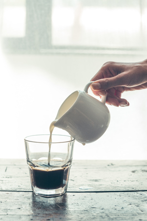 Person pouring milk from jug into glass of black coffee on wooden tableの写真素材