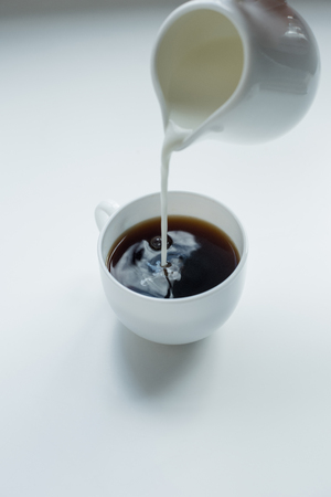 Person pouring milk from jug into cup of black coffee on white tableの写真素材