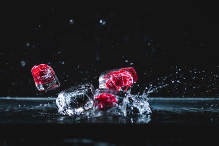 fallen ripe frozen fruits in transparent ice cubes and splashing water on blackの写真素材