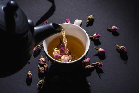 pouring tea from black kettle into cup and dry pink rose buds on black