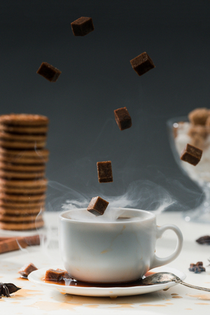 Steaming coffee cup with falling cane sugar cubes by cookies on tableの写真素材