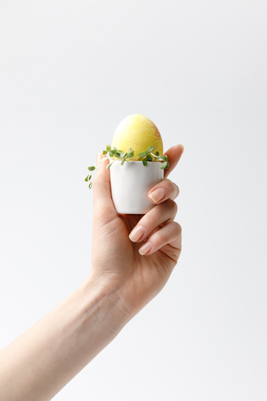 cropped image of woman holding yellow painted easter egg in egg stand isolated on whiteの写真素材