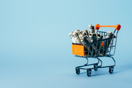 close up view of little shopping trolley with rolled dollar banknotes isolated on blueの写真素材