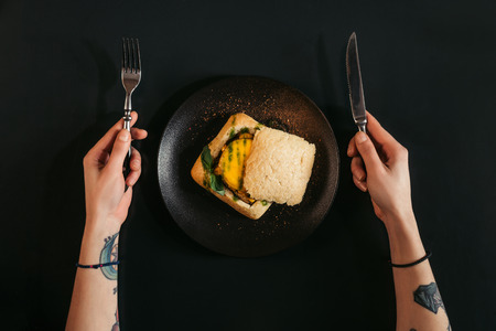 top view of person holding fork and knife while eating delicious vegan burger on blackの写真素材