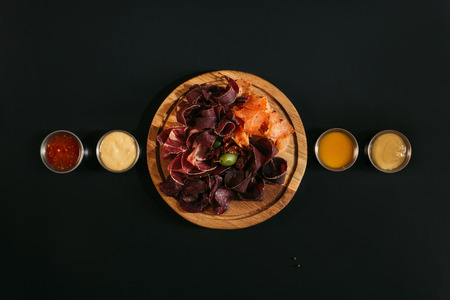 top view of delicious sliced assorted meat on wooden board with various sauces on blackの写真素材
