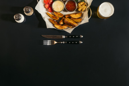top view of delicious roasted potatoes with sauces, glass of beer and fork with knife on black の写真素材