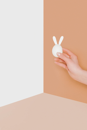 cropped view of person holding easter egg with bunny ears on beige and white minimalistic backgroundの写真素材