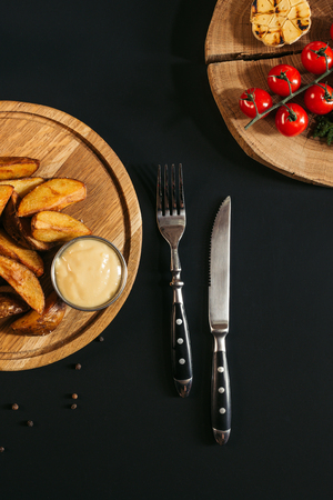 top view of baked potatoes with sauce on wooden board, fork with knife and vegetables on blackの写真素材