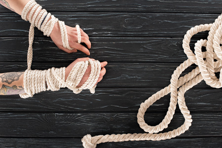 cropped shot of female tattooed hands tied in rope on dark wooden tabletop