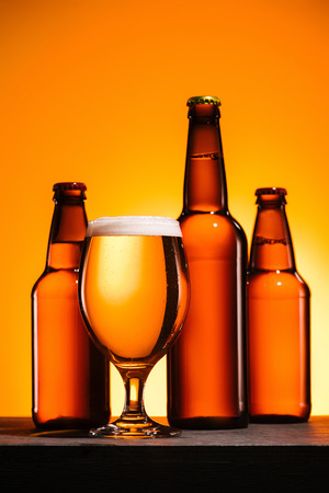 close up view of bottles and glass of beer with foam on surface on orange backgroundの写真素材