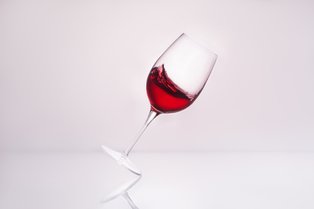 inclined wineglass with splashing red wine on reflective surface and on white