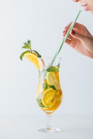 partial view of woman holding straw and summer cold cocktail with mint, lemon and orange pieces isolated on whiteの写真素材