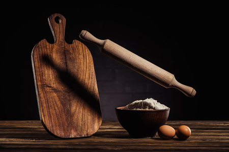 flying rolling pin, wooden cutting board, two eggs and bowl with flour on table 