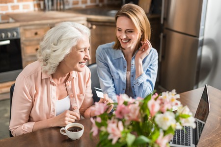 young and senior women sitting at table and using laptop in kitchenの写真素材