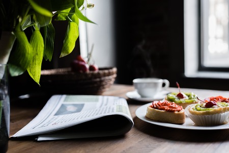 Still life of newspaper, breakfast with cakes and hot coffee on kitchen table in front of windowの素材 [FY310101977636]